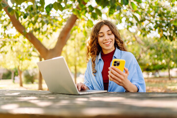 Young smiling student sit on bench in spring park outdoors, rest, use laptop, mobile cell phone...