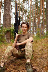 A woman in military camouflage sits on the forest floor holding a rifle amidst tall trees during a...