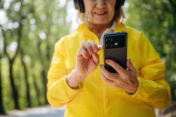 Smartphone in hands. Senior woman in yellow sport coat is outdoors with headphones