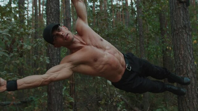 Young shirtless man is training in forest and doing one-arm hanging lateral crunches on horizontal bar.
