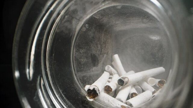 Dolly slider shot moving inside a glass jar filled with cigarette butts, capturing the texture of ashes and used filters in a confined, gritty space, emphasizing discarded items and waste.