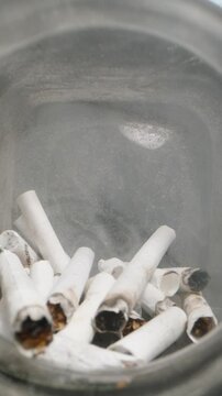 Vertical video. Dolly slider, close up. View through the mouth of a glass jar filled with cigarette butts, showing used filters and ash inside, emphasizing waste, pollution, and the impact of smoking 