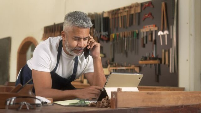Focused craftsman multitasking in a workshop. Mature man talking on the phone while taking notes and using a tablet, showcasing small business management.