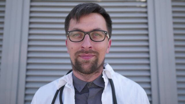 Young male doctor with glasses and stubble, dressed in white coat and protective mask, stands outdoors, removing his mask with confident smirk. Healthcare professional. Portrait of physician intern. 