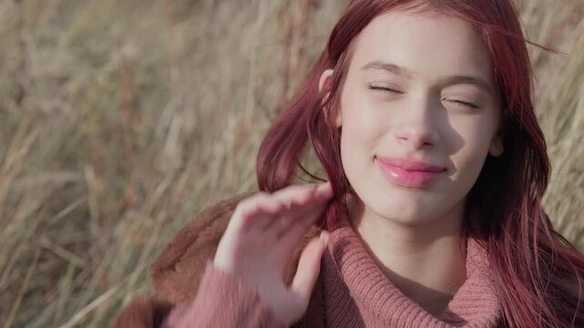 Teen girl smiling shyly while adjusting her hair, dressed warmly in a cozy autumn landscape.