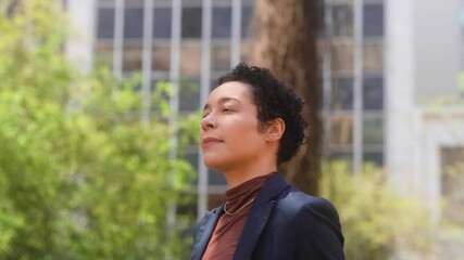 Confident African American woman with short curly hair walking in urban setting surrounded by trees. Professional individual dressed in business attire navigating city landscape
