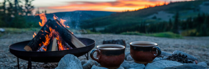 A banner depicting a campsite with a campfire and two ceramic cups filled with hot tea. The photo shows a bonfire in which logs are burning. A mountain landscape is depicted in the background.