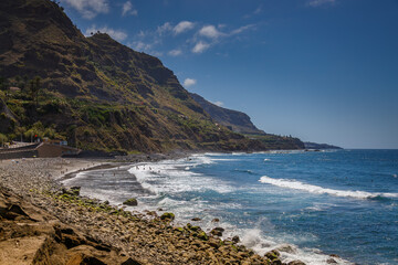 Obraz premium Tenerife's El Socorro beach with powerful ocean waves meeting rocky sands, green cliffs in the distance
