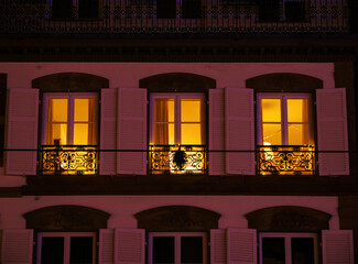 Warm yellow light glows from the windows of a charming building, revealing an inviting atmosphere. The ornate ironwork balcony adds character to the nighttime scene