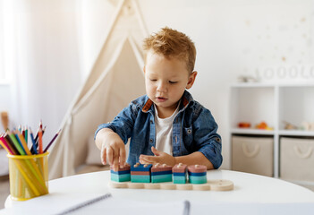 Adorable little toddler boy sits at a round table, carefully stacking colorful blocks. The room is well-lit and inviting, filled with toys and art supplies.