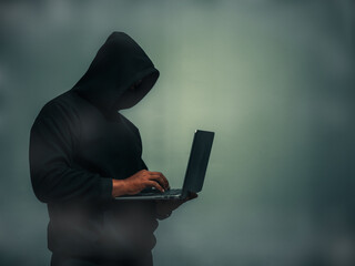 Faceless anonymous hacker in black hoodie and covering his face holding a laptop with a light...