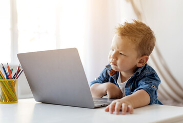 Portrait of little curious boy examines the screen of his laptop, seated at a cluttered desk with art supplies, illuminated by natural light streaming through a window.