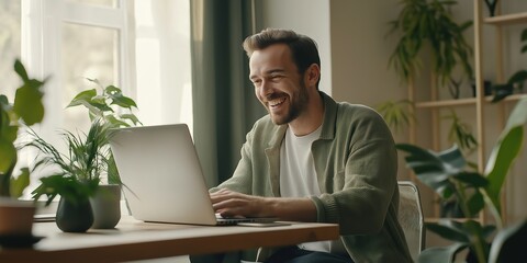 A man is sitting at a desk with a laptop in front of him. He is smiling and he is enjoying himself. The room has several potted plants, including one on the left side of the desk