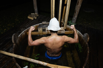 Man in wooden sauna tub flexing arms with towel hat outdoors