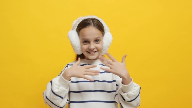 Smiling cheerful little girl wearing striped shirt and fur headphones beckoning with her hands inviting someone to come here with playful gesture isolated on yellow background