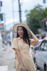 A woman wearing a straw hat and a tan dress is smiling and holding a handbag