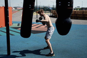 Young muscular man on boxing training, hitting punching bag, workout outside on sports ground