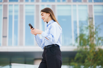 Young Woman Using Smartphone While Walking Outside a Modern Building on a Sunny Day in an Urban Area