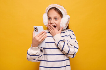 Little girl scrolling through smartphone browsing internet surprised by content shocked by new information addicted to technology absorbed in social media isolated on yellow background