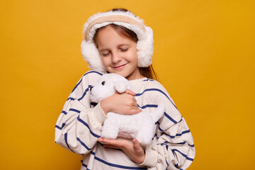 Smiling female child receiving stuffed dog as present embracing it with closed eyes and charming smile feeling happy while playing with toy indoors isolated over yellow background