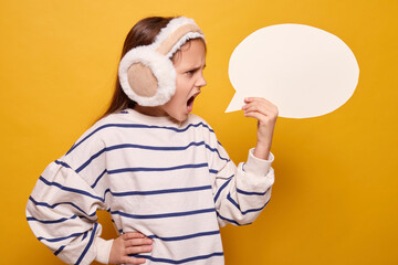 Screaming schoolgirl with fur headphones holding empty speech bubble exclaiming angrily informing about something offering free space for idea in advertisement mockup isolated over yellow background