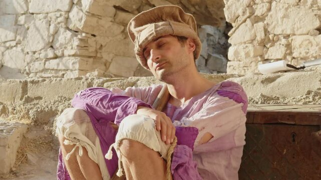 An adult young man of Arab appearance in a medieval old costume sleeps while sitting on the ruins