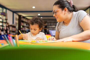 Latina teacher smiling as she guides her preschool student in an art project where the child is...