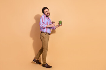 Fototapeta premium Full size photo of nice young male walk phone call hold coffee wear trendy striped formalwear isolated on beige color background