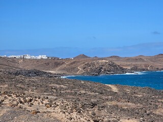 Un paysage aride et rocailleux à Costa Teguise, avec des collines de pierres volcaniques brunes qui s'étendent jusqu'à l'horizon. Des maisons blanches, typiques de l'architecture locale, sont visibles