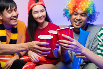 Fototapeta premium A group of excited Asian male and female friends watched the World Cup match on TV and drank beers and ate snacks together at home. Sports fans shout and celebrate the victory of their sports team.