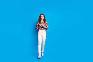 Full length photo of lovely young lady hold gadget walking dressed stylish colorful garment isolated on blue color background