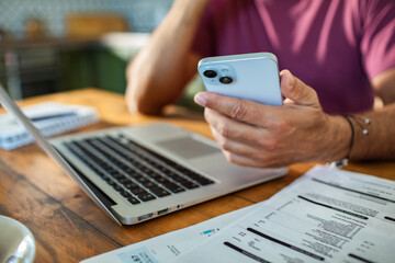 Man checking phone while reviewing bills and using laptop at kitchen table