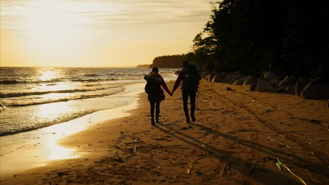 Love and travel, hikers holding hands and strolling together on coast in sunset. Back view of two happy people walking on sea shore and carrying backpacks, local tourism in nature, slow motion shot