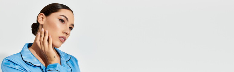 A young woman in a chic blue outfit touches her face, radiating elegance and thoughtfulness.