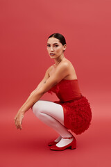 A young beautiful woman crouches gracefully, showcasing her red outfit and striking expression.