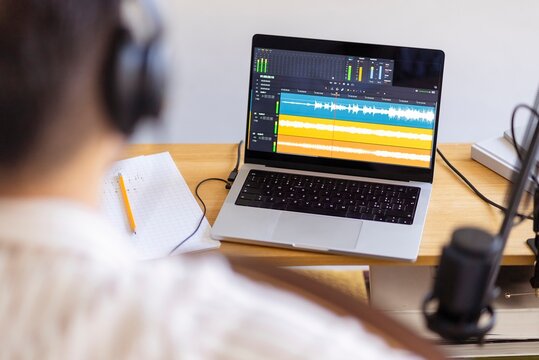 A musician is captured recording music in a home studio setup, with a microphone and a laptop displaying sound editing software