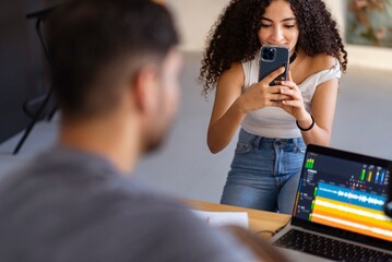A young woman holds her smartphone up to photograph a man working with audio editing software on his laptop, displaying sound waveforms and project details for an ongoing recording