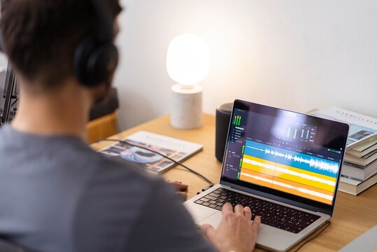 A man wearing headphones works on audio editing software on his laptop, focusing on the sound waveforms displayed on the screen as he adjusts settings in a calm workspace lit by a small desk lamp
