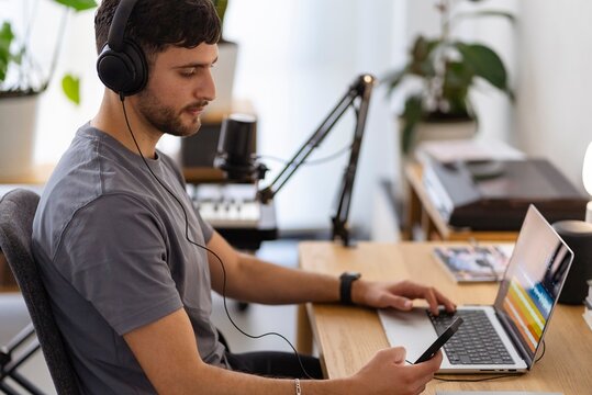 A man wearing headphones operates audio editing software on his laptop while holding a smartphone, with a professional microphone nearby, in a well-lit workspace with plants and recording equipment