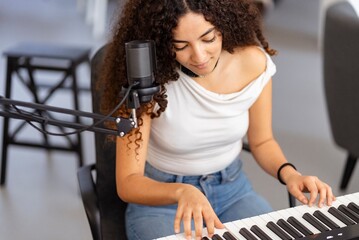 A woman with curly hair focuses on playing an electronic keyboard in a home studio setup, with a microphone positioned nearby for recording, indicating a dedication to musical practice and creativity