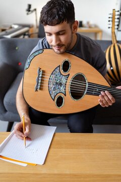 A man holds a traditional oud instrument while writing musical notes on paper, seated in a home studio environment