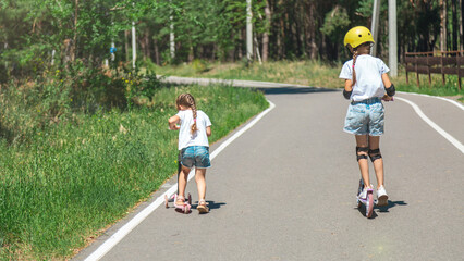 Caucasian children girls together in sunny weather in summer park riding on kick scooters. Happy...