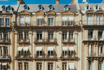 Close-up of classic Parisian building facade with ornate balconies, large windows, and detailed architecture under a bright blue sky, showcasing elegance and urban charm.

