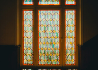 Colorful stained glass window with geometric patterns and vibrant red, yellow, and green tones, illuminated by natural light, showcasing intricate craftsmanship and artistic design.