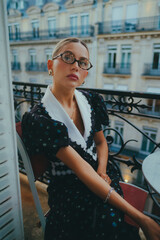 Elegant woman with glasses sitting outdoors, wearing a vintage floral dress with lace details, against a backdrop of classic European architecture, showcasing beauty and sophistication.