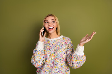 Cheerful young woman in colorful sweater chatting on phone, expressing delight with raised hand...