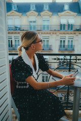 Fototapeta premium Elegant woman with glasses sitting outdoors, wearing a vintage floral dress with lace details, against a backdrop of classic European architecture, showcasing beauty and sophistication.