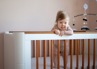 Portrait of a cute Caucasian baby girl 6 months old standing in a crib and looking at the camera. Copy space