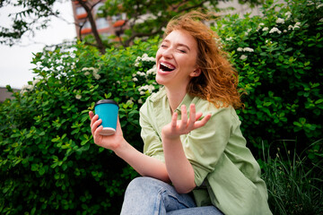Fototapeta premium Joyful young redhaired woman enjoying a sunny day outdoors holding coffee and laughing in natural light