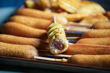 Pan full of Corn dogs, one with mustard and fries in background. Selective focus with blurred...